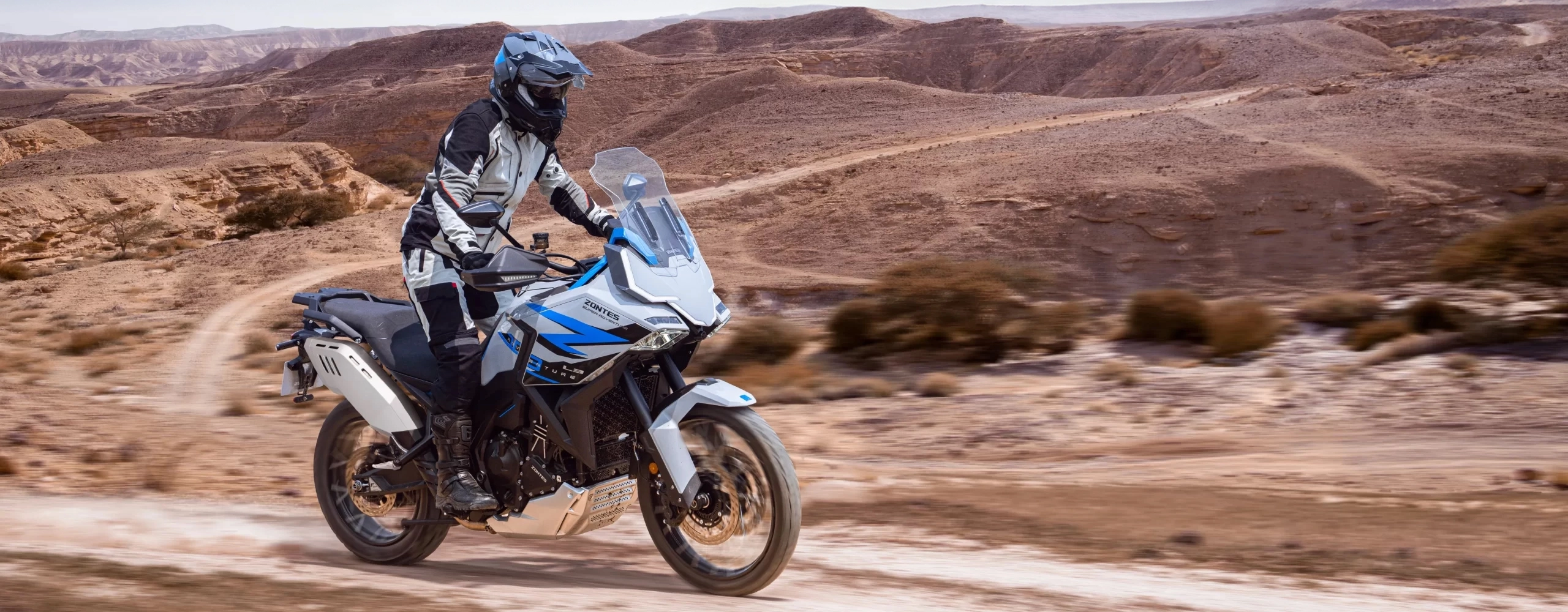 Zontes motos en Uruguay - Moto Zontes 703F sobre terreno seco con piloto de aventura y fondo de cerros. Zontes motos en Uruguay - Zontes 703F circulando por un camino de tierra en un paisaje árido con conductor equipado.
