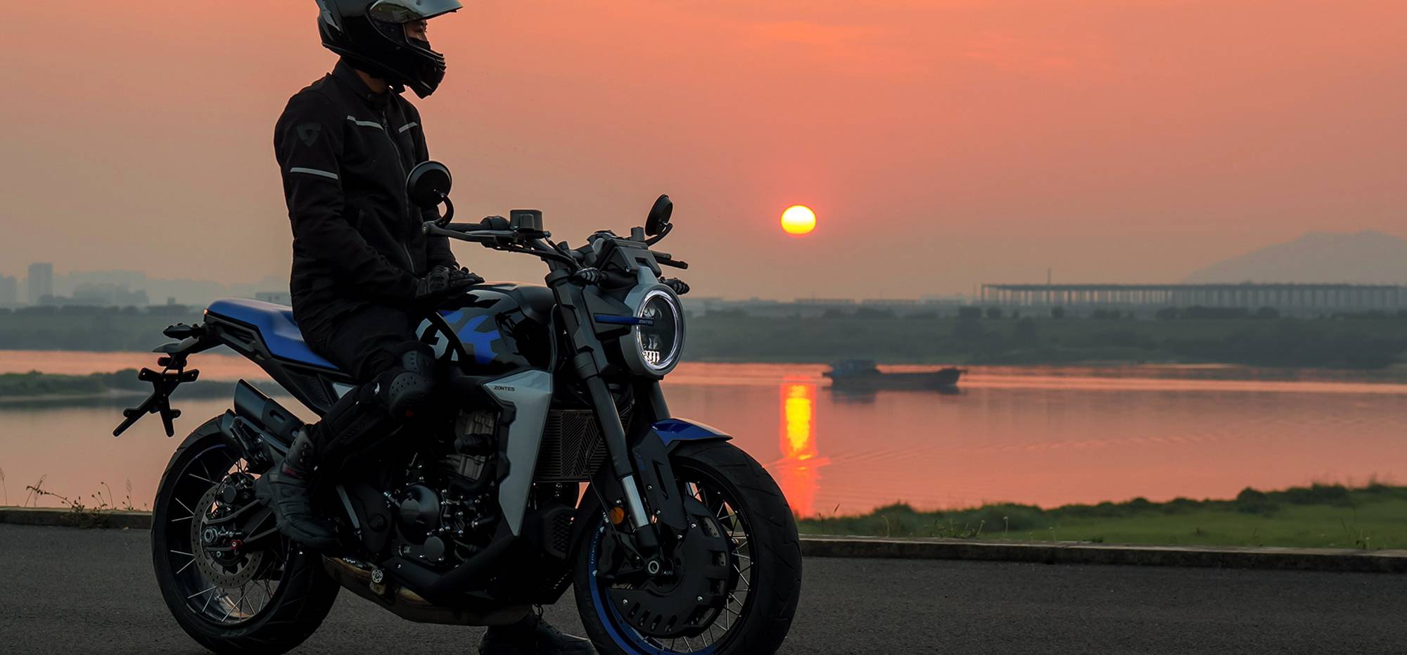 Zontes motos en Uruguay - Moto Zontes 350GK junto a un piloto detenido en una escena exterior al atardecer. Zontes motos en Uruguay - Zontes 350GK detenida con conductor equipado frente al agua y una puesta de sol al fondo.