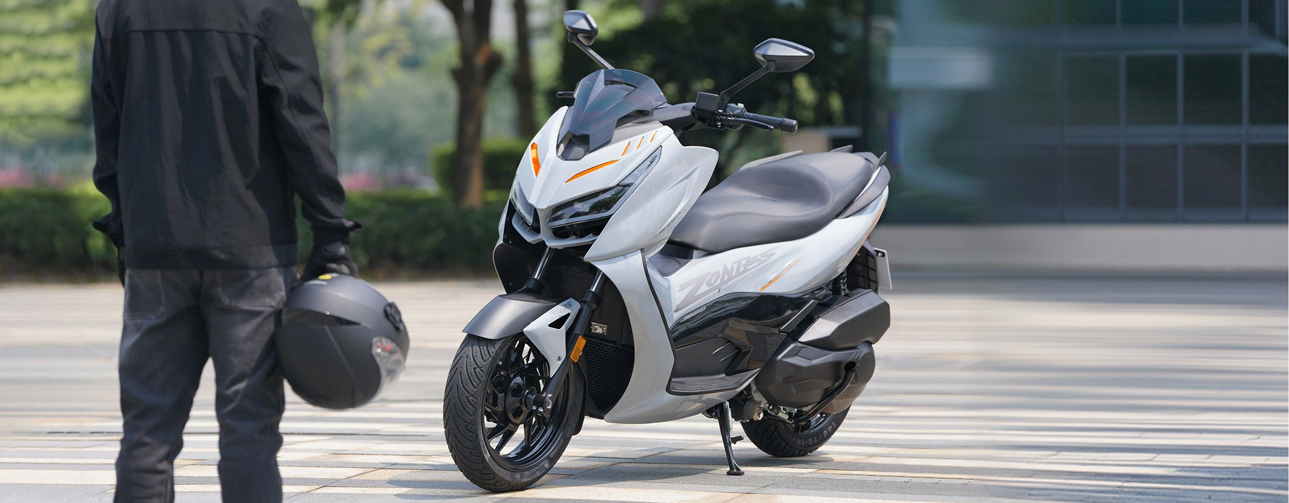 Zontes motos en Uruguay - Scooter 368D estacionado con pata lateral desplegada y acompanamiento de conductor equipado. Zontes motos en Uruguay - Zontes 368D de carroceria clara detenido sobre una explanada junto a una persona con casco en la mano.